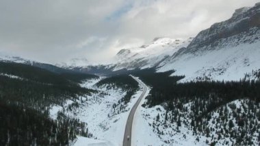 Kanada Alberta 'da orman ve karla kaplı dağlarla çevrili bir şekilde yol boyunca giden aracın insansız hava aracı görüntüleri.