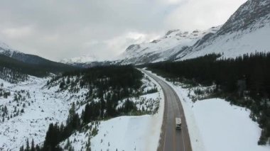 Kanada 'nın Alberta kentindeki karlı dağlar ve kozalaklı ormanlar arasında yol boyunca ilerleyen karavanın hava manzarası.