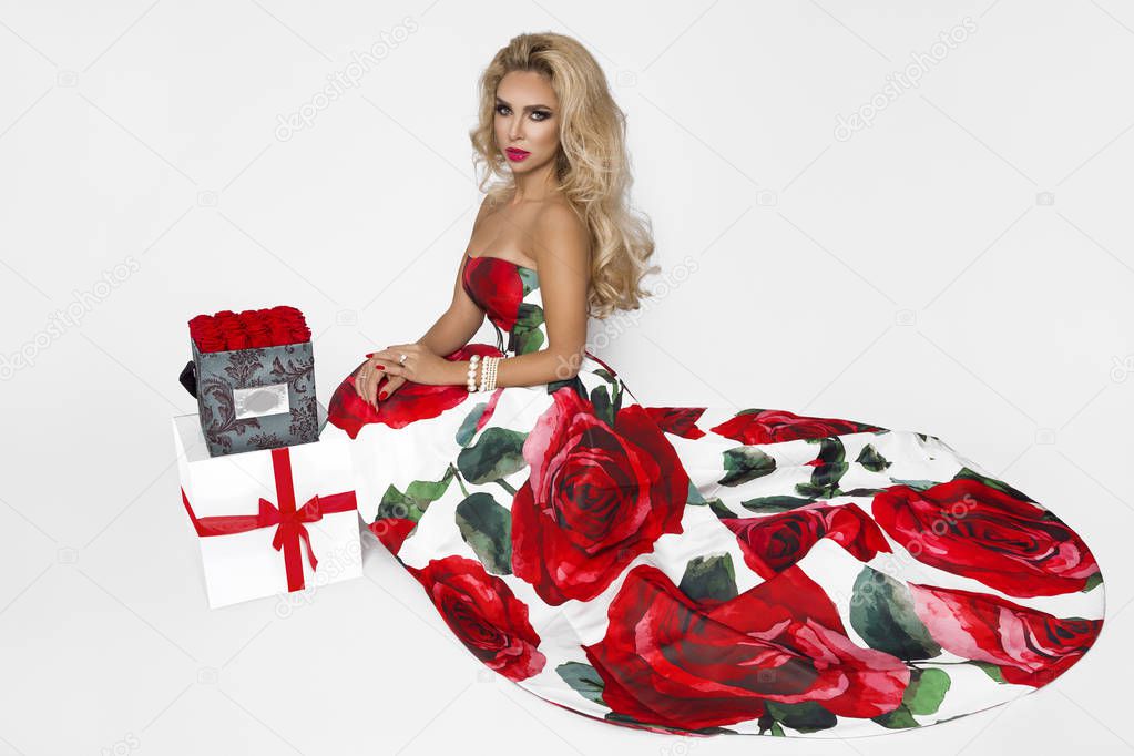 Beautiful blonde woman in an elegant evening gown with red roses, holding a present. Chrismas time. A model posing in the studio on a white background.