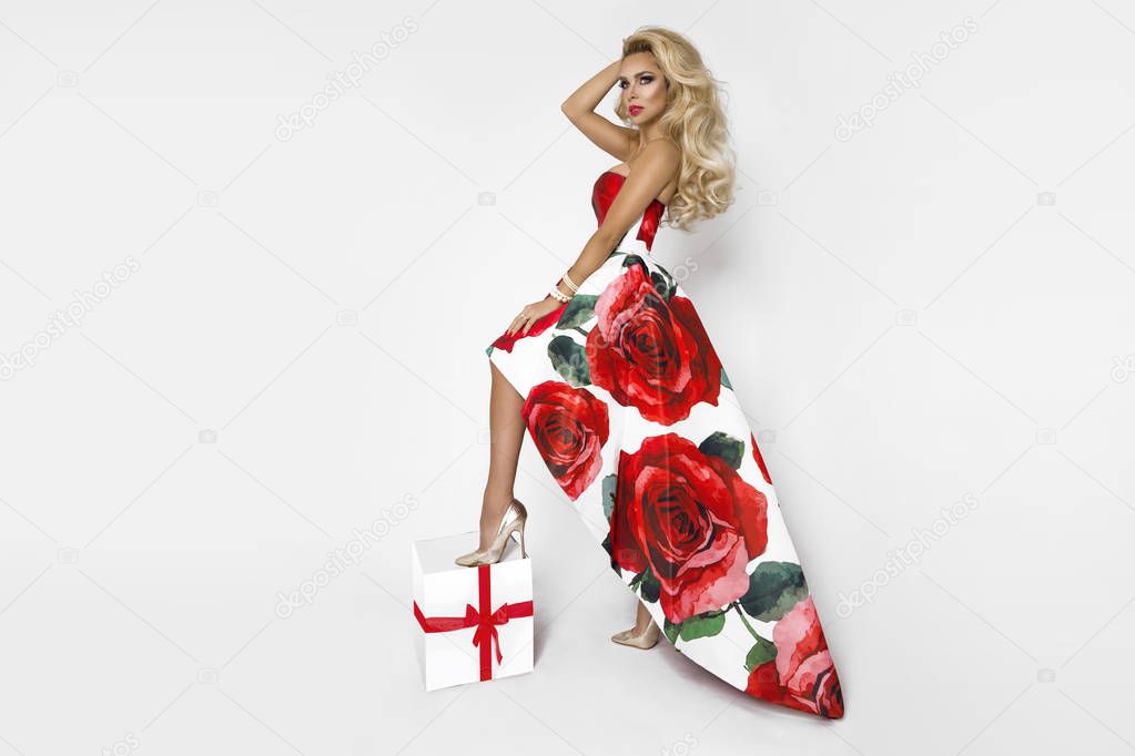 Beautiful blonde woman in an elegant evening gown with red roses, holding a present. Chrismas time. A model posing in the studio on a white background.