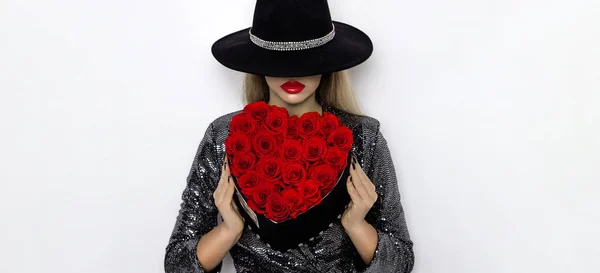 Valentine Beauty girl with red heart roses. Portrait of a young female model with gift and hat, isolated on background. Beautiful Happy Young woman presenting flower box. Holiday party, birthday.
