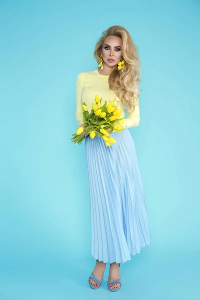 Spring Woman. Beauty Summer model girl with colorful clothes, holding a bouquet of spring flowers. Beautiful lady with yellow tulips on the blue background.