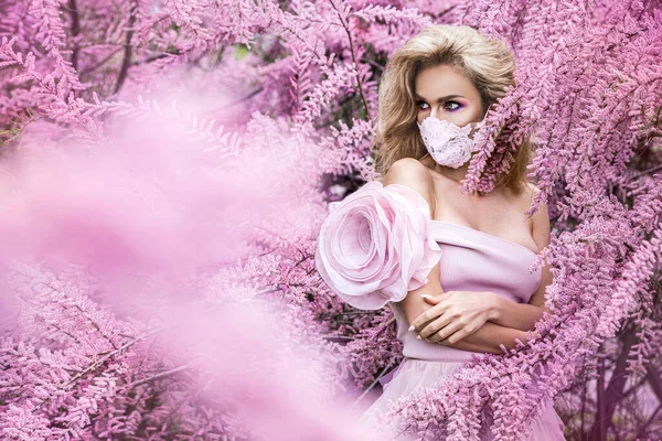 Fashion portrait of young woman wearing a face mask and pink clothes, in a flowering garden.Pandemic, virus, coronavirus, Covid-19 masked girl. Spring and summer fashion, model in a mask.