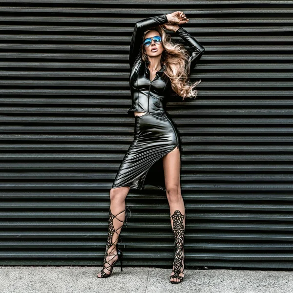 Modaya uygun. Siyah lateks kıyafetler ve siyah arka planda izole edilmiş güneş gözlüğü takan alımlı güzel bir model. Seksi, şık, sarışın kadın. Deri ve lateks kavramı. Kaya stili.