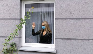 Coronavirus. Corona virüsünün hasta kadını pencereden dışarı bakıyor ve maske takıyor ve evdeki hastalıktan korunuyor. Kilitleme kavramı.