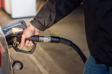 Mavi kot pantolonlu ve siyah ceketli bir adam benzin dolduran pompa tutuyor. Tanka benzin pompalıyor. Benzin istasyonunda bir araba yakıt ikmali