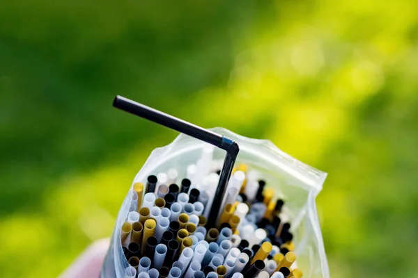 White, black and gold plastic straws in a plastic bag on green grass background. Plastic pollution, environmental waste. Say no to plastic straw