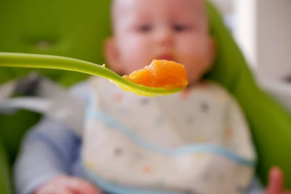 Colher cheia de cenoura pura e um bebê embaçado no fundo. Foco seletivo na colher. Bebê primeiro soilds. Desmame . — Fotografia de Stock