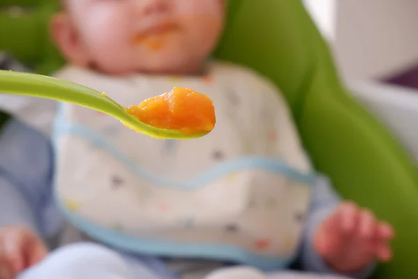 Colher cheia de cenoura pura e um bebê embaçado no fundo. Foco seletivo na colher. Bebê primeiro soilds. Desmame . — Fotografia de Stock