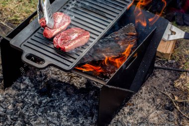 Biftekler kamp ateşinde dökme demir tabakta ızgara yapıyor. Kamp ateşi pişirme. Açık hava mangalı