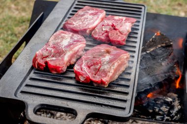 Biftekler kamp ateşinde dökme demir tabakta ızgara yapıyor. Kamp ateşi pişirme. Açık hava mangalı