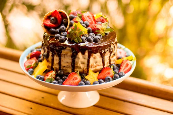 Delicious handmade layered cake decorated with chocolate glaze, strawberries, blueberries, carambola star fruit and ice cream cone on nature tropical background.