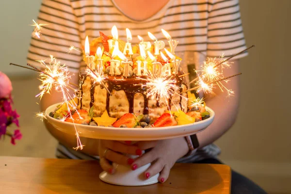 Delicious handmade layered cake decorated with sparkles, happy birthday candles, chocolate glaze, strawberries, blueberries, carambola star fruit and ice cream cone on nature tropical background.