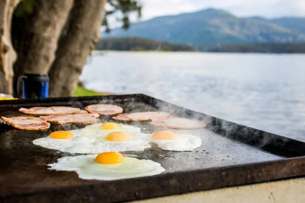 Breakfast camp cooking, Grilling eggs and bacon on the bbq plate with beautiful nature landscape on background.