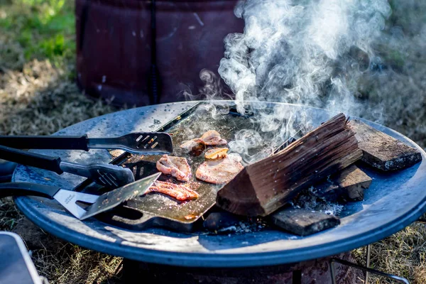Breakfast camp cooking. grilling crispy bacon on a cast iron plate over the camp fire. Selective focus.