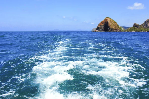 Trail and foamy waves on blue sea surface behind of moving boat with island background. — Stock Photo, Image