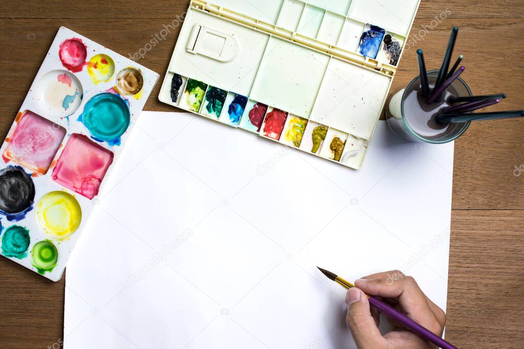 Man hand holding paint brush on white paper with colorful watercolor and equipment on wooden background.