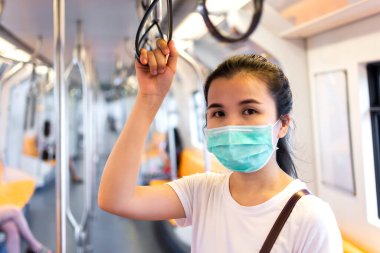 Tıbbi hijyen koruyucu yüz maskesi takan genç Asyalı kadın yolcu büyük şehirde metro ya da gökyüzü treninde duruyor ve tutuş halkası tutuyor..