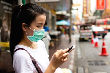 Tıbbi hijyen koruyucu yüz maskesi takan genç Asyalı kadın gezgin büyük şehirde akıllı telefon kullanıyor..