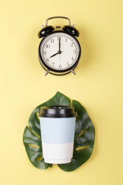 Canavar yaprağının üzerinde kağıt kahve fincanı ve sarı renkli arka planda alarm saati. Günaydın uyan konsepti. Düz üst görünüm 