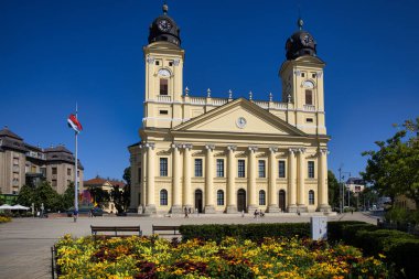 Debrecen, Hungary July 30, 2025: Great Reformed Church with twin towers and neoclassical facade. Colorful summer flowers bloom in foreground with Hungarian flag against clear blue sky