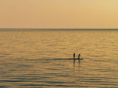 Samothraki, Greece  sup activity at Kipos Beach during sunset, with two people silhouetted on a paddleboard over calm waters, golden reflections, and distant hills in a serene coastal setting.