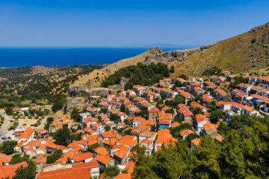 Açık bir yaz gökyüzünün altında, derin mavi denizin üzerindeki yeşil bir yamaçta, kırmızı çatıları ve şatosu olan geleneksel köy..