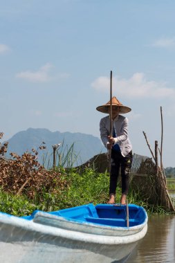Hpa-An, Myanmar - 19 Kasım 2018: Geleneksel giysi ve şapka giyen genç birmanyalı kadının dikey resmi, Hpa-An'daki Sadan Mağarası'na yakın nehirde yelkenli tekne,
