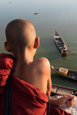 Mandalay, Myanmar'da bulunan U Bein Köprüsü'nden teknelere bakan genç Budist keşişin dikey resmi