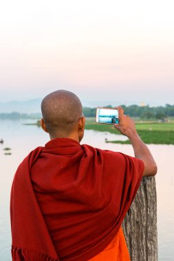 Mandalay, Myanmar - 03 Aralık 2018: Mandalay, Myanmar'da gün batımında U Bein Köprüsü'nden nehrin fotoğraflarını çeken Budist keşişin dikey fotoğrafı
