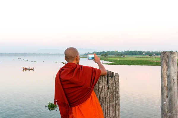 MANDALAY, MYANMAR - 03 DECEMBER, 2018: Horizontal picture of buddhist monk taking pictures of the river from U Bein Bridge during sunset in Mandalay, Myanmar