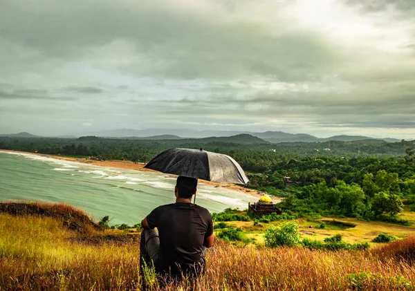 man sitting on hilltop with umbrella enjoying rain with amazing view image is taken at gokarna karnataka india. the view from here is pristine and mesmerizing.