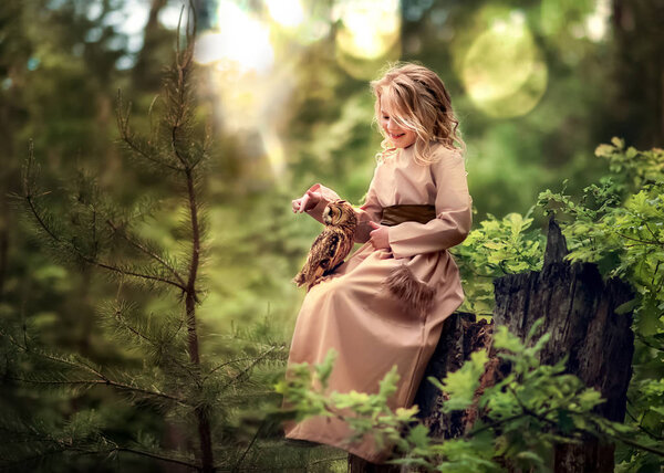 A little beautiful girl with blond long hair sits in the green forest on a high stump in the summer and plays with a live owl.
