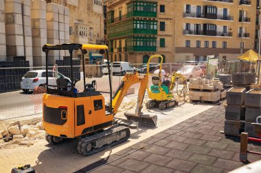 Kentsel yol onarımı: arka planda taş bloklar, eskrim ve şehir manzarası olan bir inşaat alanında kompakt kazıcılar