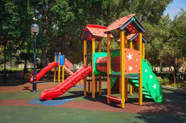 Slaytlı renkli oyun alanı ve ağaçların arasındaki güneşli bir parkta yumuşak kauçuk zeminde yapılar..