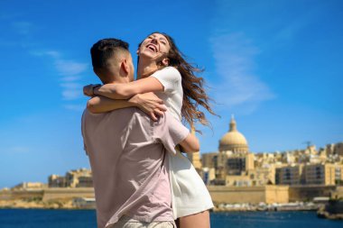 Arka planda tarihi şehir ve Akdeniz ile kucaklaşan mutlu genç çift. Kameraya bakan adam, uzun siyah saçlı kadın neşeyle gülüyor..