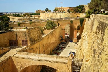 Malta Valletta 'daki antik taş tahkimatları ve kemerli köprülerin panoramik manzarası. Kale duvarları ve şehir manzarası yakınındaki park halindeki arabalar..