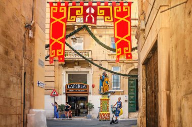 Valletta, Malta - 09.17.2025: Şenlikli süslemeleri olan şehir caddesi, yol kesişiminde bir azizin heykeli ve yerel halk.