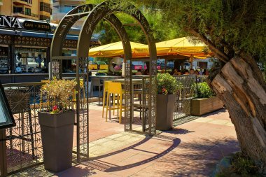 Sliema, Malta - 07.06.2025: Girişte metal kemer ve saksı bitkileri olan bir ağacın altında rahat bir kafeterya. İnsanlar güneşli bir günde sarı şemsiyelerin altında terasta dinlenirler..