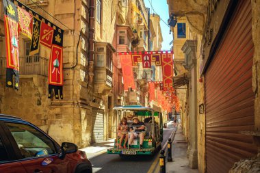 Valletta, Malta - 09.17.2025: Turistler, katolik festivali için renkli bayraklarla süslenmiş dar, eski bir caddede üstü açık bir gezici arabayla geziyorlar.