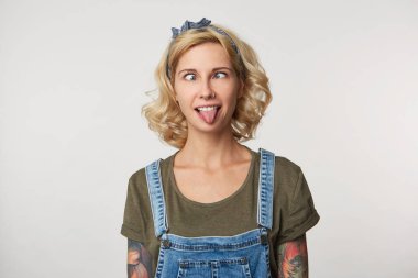 Indoor shot of young female has fun, makes grimace and shows tongue, weras casual denim overalls and t shirt. Portrait of funny young woman being crazy clipart