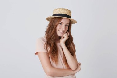 Kızıl saçlı Genç kadın hasır şapka ve katlanmış elleriyle ayakta tshirt büyüleyici ve beyaz arka plan üzerinde gülümseyen mutlu portre