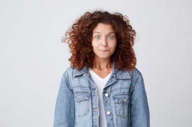 Denim ceket yapar komik surat ve doğrudan fotoğraf makinesindeki beyaz arka plan üzerinde izole görünüyor eğlenceli çok kıvırcık saçlı Genç kadın portresi giyer