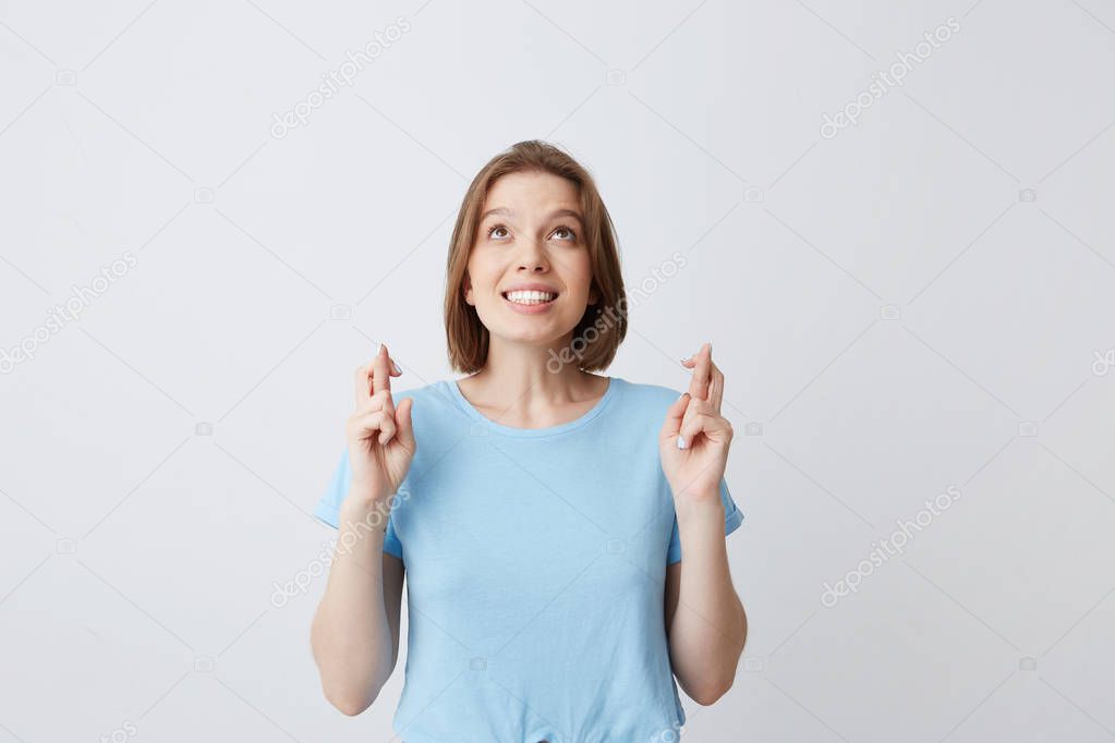 Cheerful pretty young woman in blue tshirt with fingers crossed looking up and wishing for happiness isolated over white background Feels happy and inspired