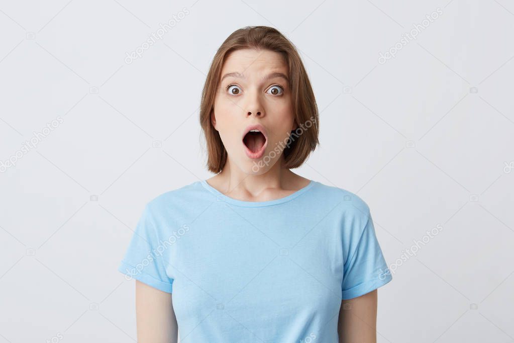 Closeup of shocked stunned young woman in blue t shirt with opened mouth feels amazed and looks directly in camera isolated over white background