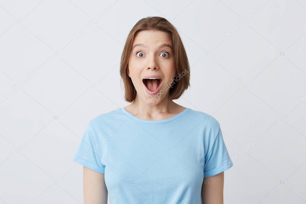 Portrait of surprised happy young woman with opened mouth in blue t shirt feels excited and shouting isolated over white background