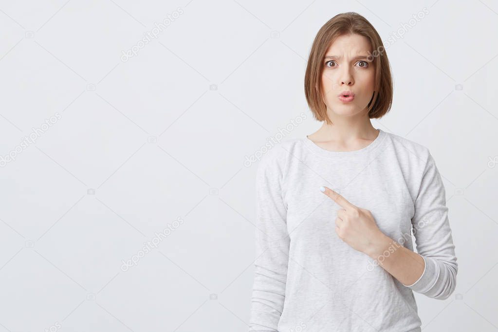 Portrait of astonished pretty young woman in longsleeve feeling amazed and pointing to the side on copyspace isolated over white background