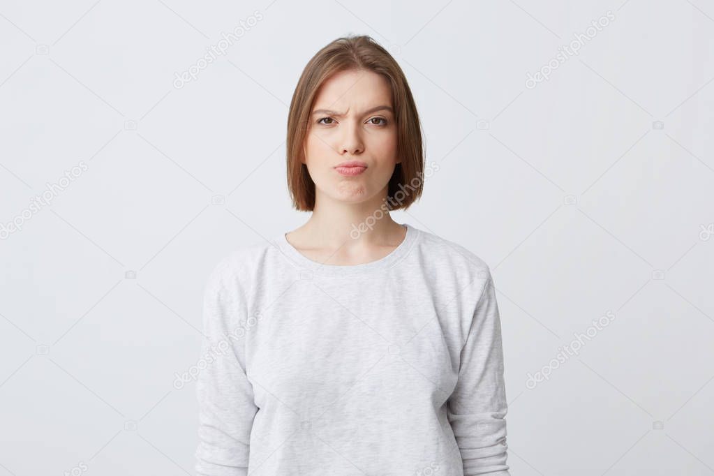 Portrait of serious unhappy young woman in longsleeve standing and frowning her face isolated over white background Looking directly in camera