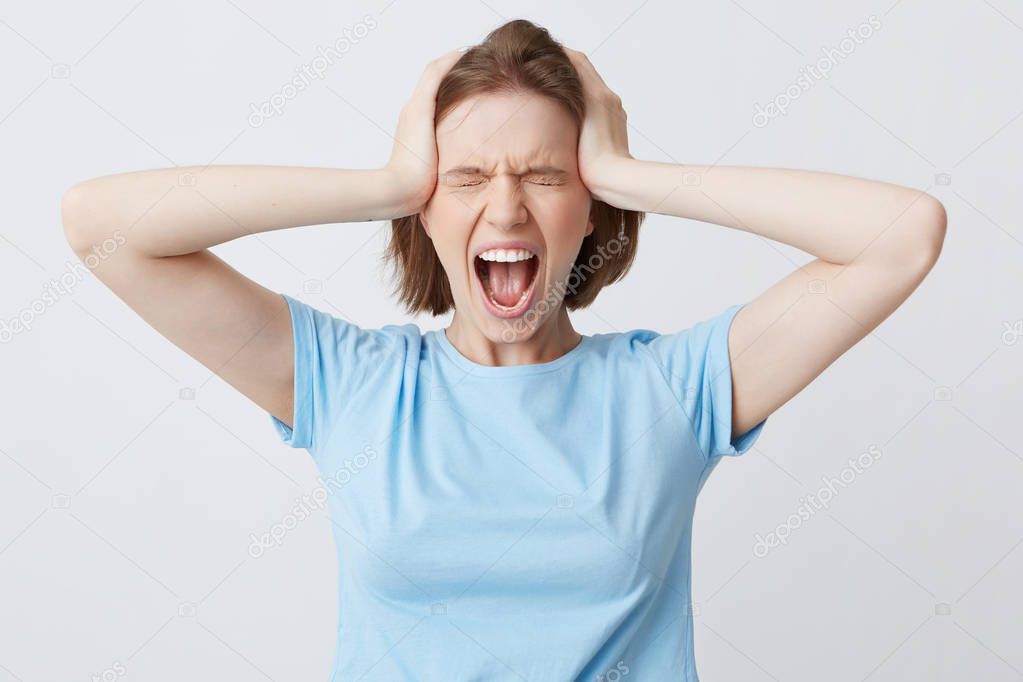 Portrait of crazy hysterical young woman in blue tshirt with closed eyes and hands on head looks stressed and screaming isolated over white background