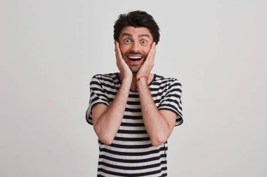 Indoor shot of optimistic male with glad expression, keeps hands on cheeks, feels shoked and happy, isolated over white wall clipart